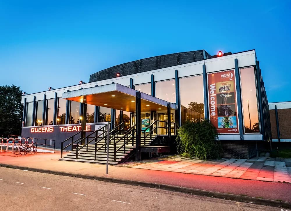 The Queen's Theatre building at dusk, featuring a modern entrance with steps and a red banner advertising upcoming shows.
