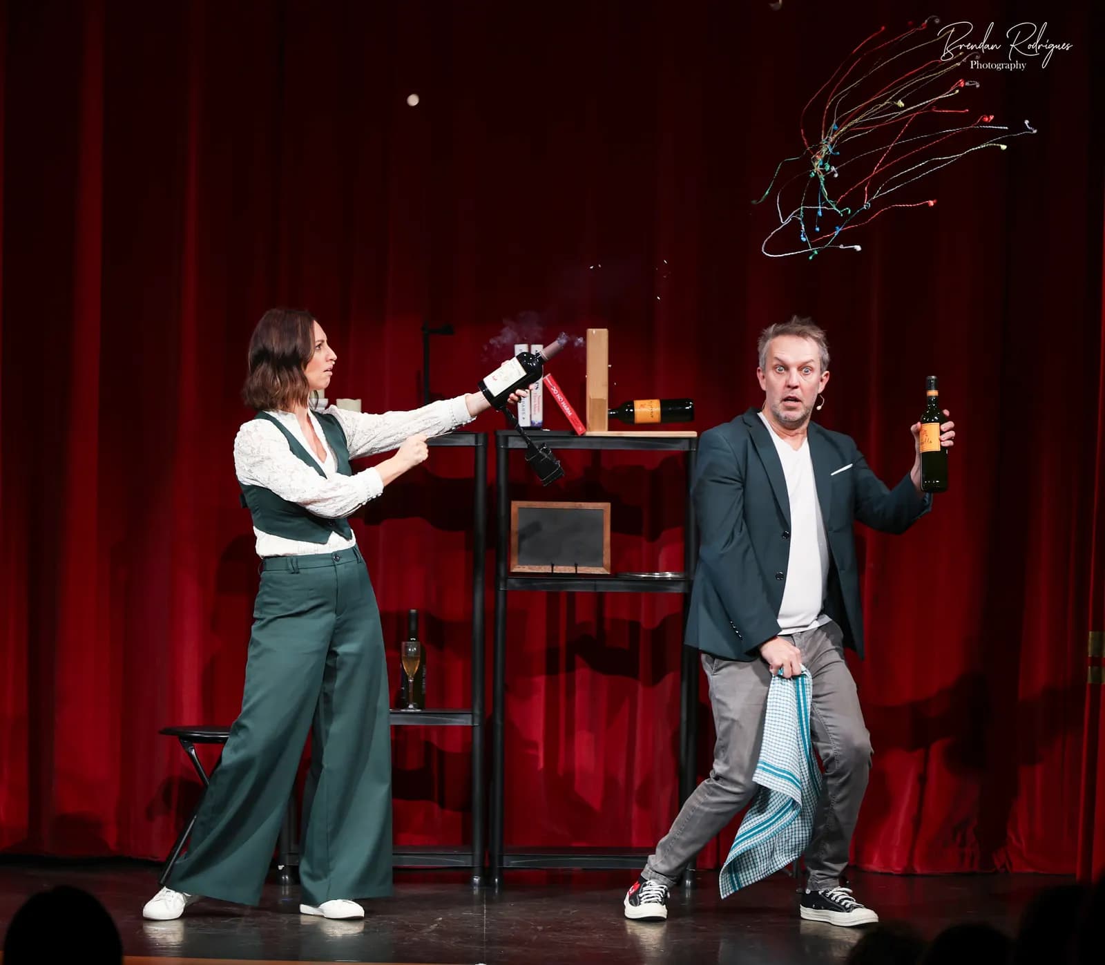 A woman aims a smoking wine bottle as a man holding another bottle looks surprised, with red curtains and a shelf in the background.