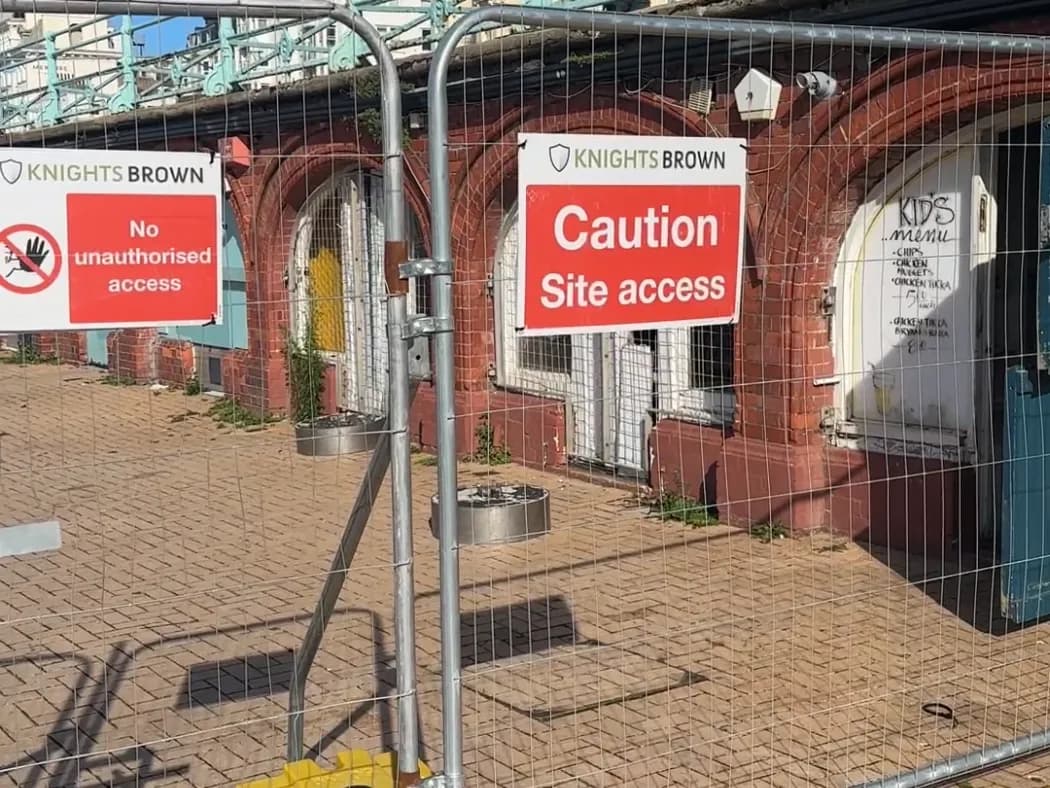 A chain-link fence blocks access to a brick building with arched doorways, two red "KNIGHTS BROWN Caution Site access" signs are visible.