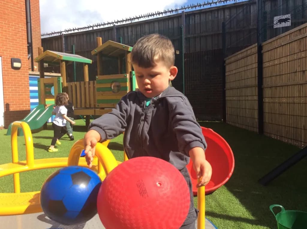 A young boy in a grey hoodie stands on a green turf playground, looking down at two balls on a yellow structure in front of him, with other children and playground equipment in the background.