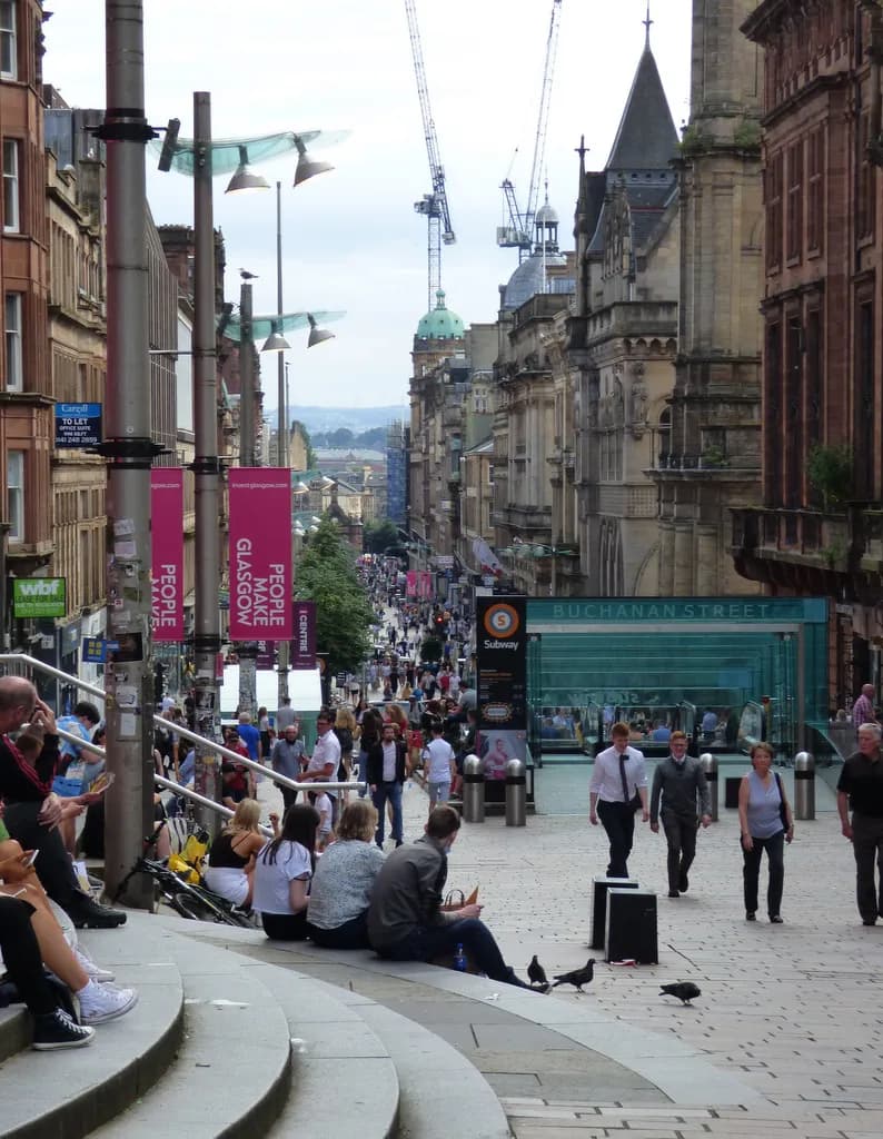 A busy street scene in a city, with people walking and sitting on steps, and tall buildings lining the street under a cloudy sky.