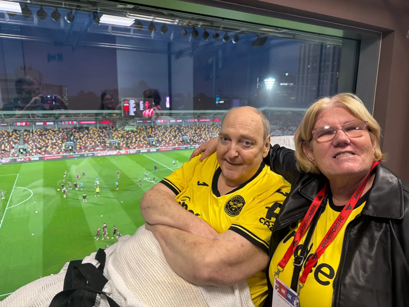 Two older people in yellow shirts, sitting in a stadium box and smiling at the camera, with a football match happening below them.