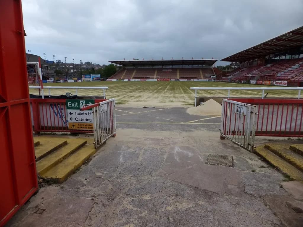 A view into an empty football stadium with red seating and a patchy green field, seen through an open gate.