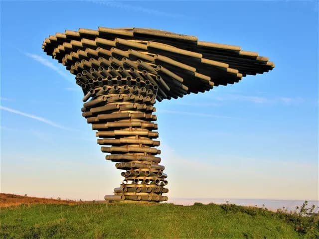 A large, twisting metal structure made of stacked pipes, resembling a tree or funnel, stands on a grassy hill against a blue sky.