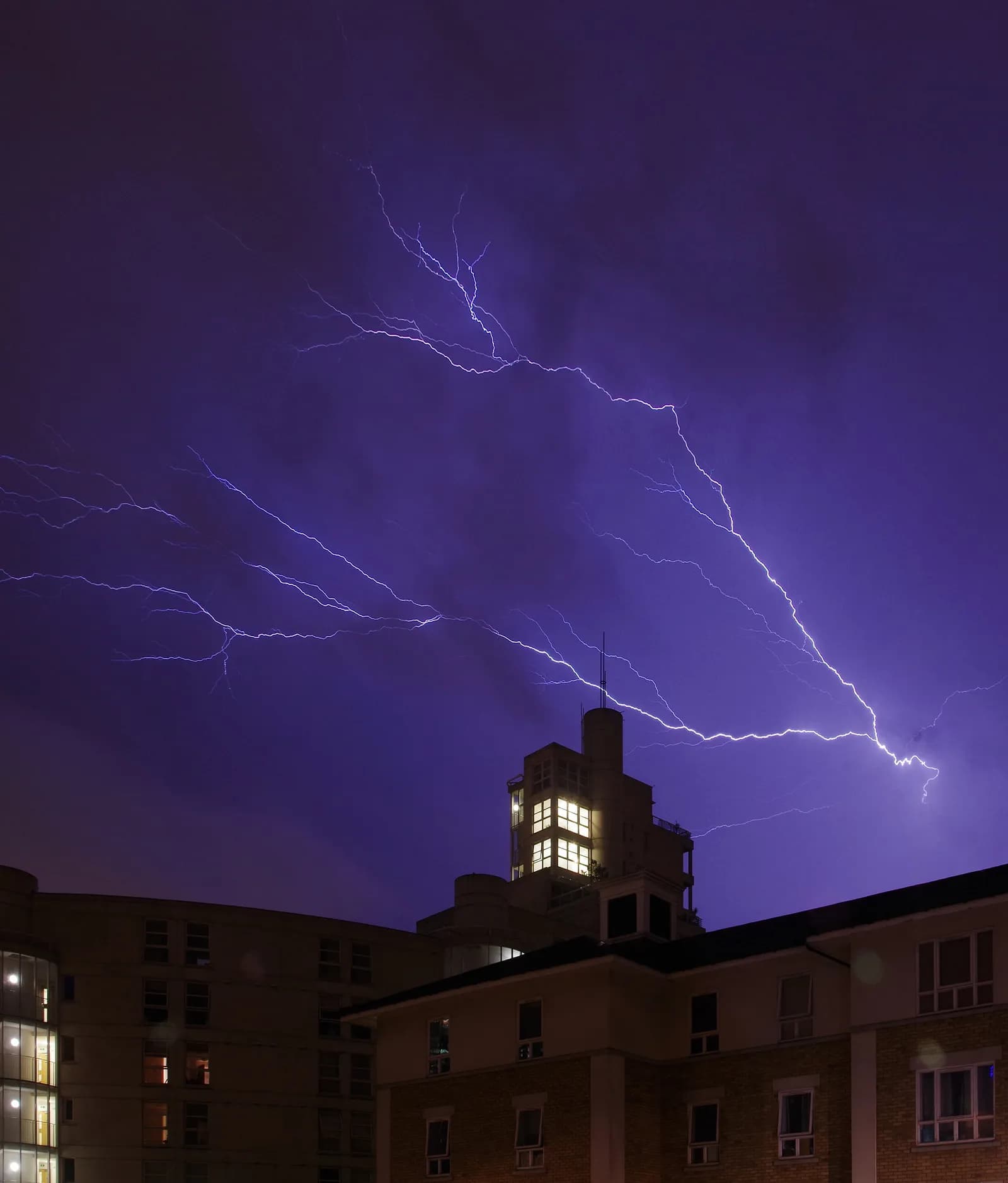 Lightning strikes illuminate a dark purple sky above several buildings at night, with one building having lit windows.