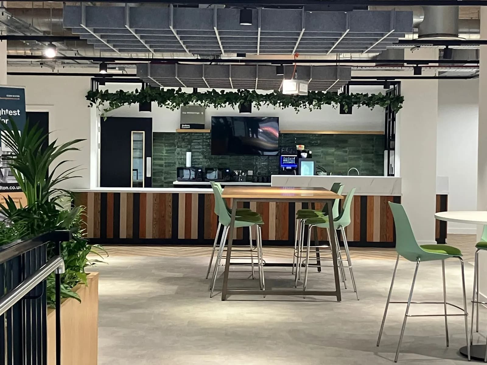 A modern office breakroom features a long counter with a wooden and white striped facade, green tiled backsplash, and various appliances, with a TV screen mounted above, while a wooden table with green bar stools occupies the foreground.