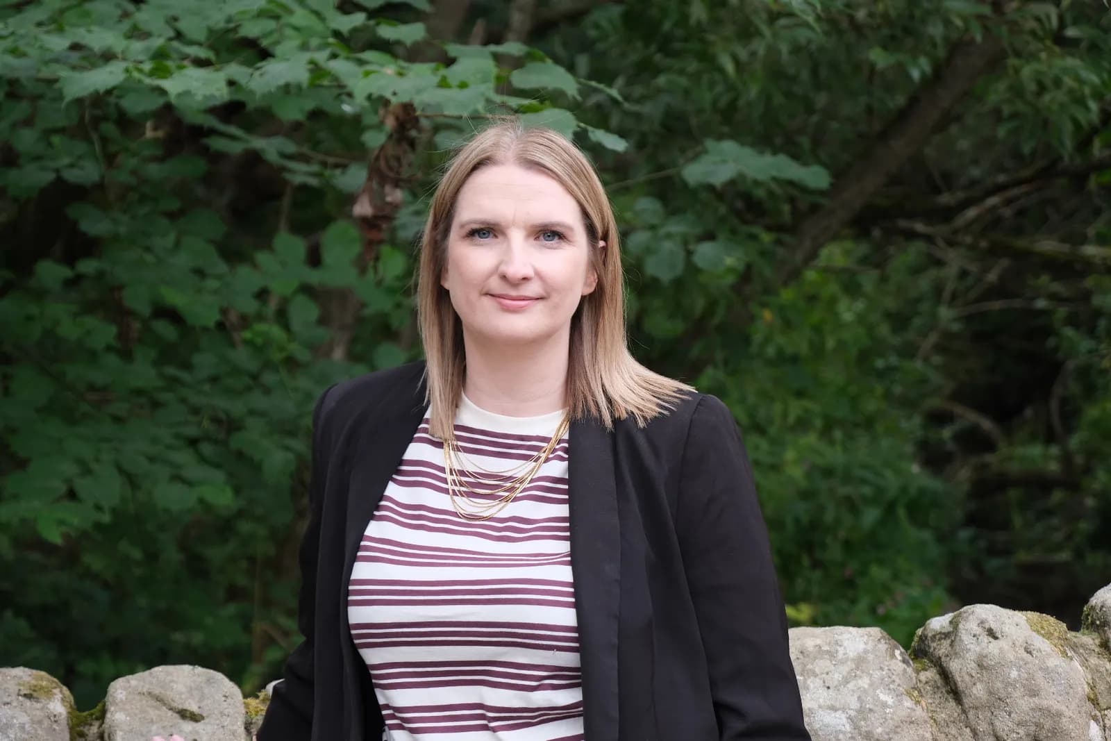 A woman with blonde hair and blue eyes, wearing a striped shirt and black blazer, standing in front of a leafy green background with a stone wall in the foreground.