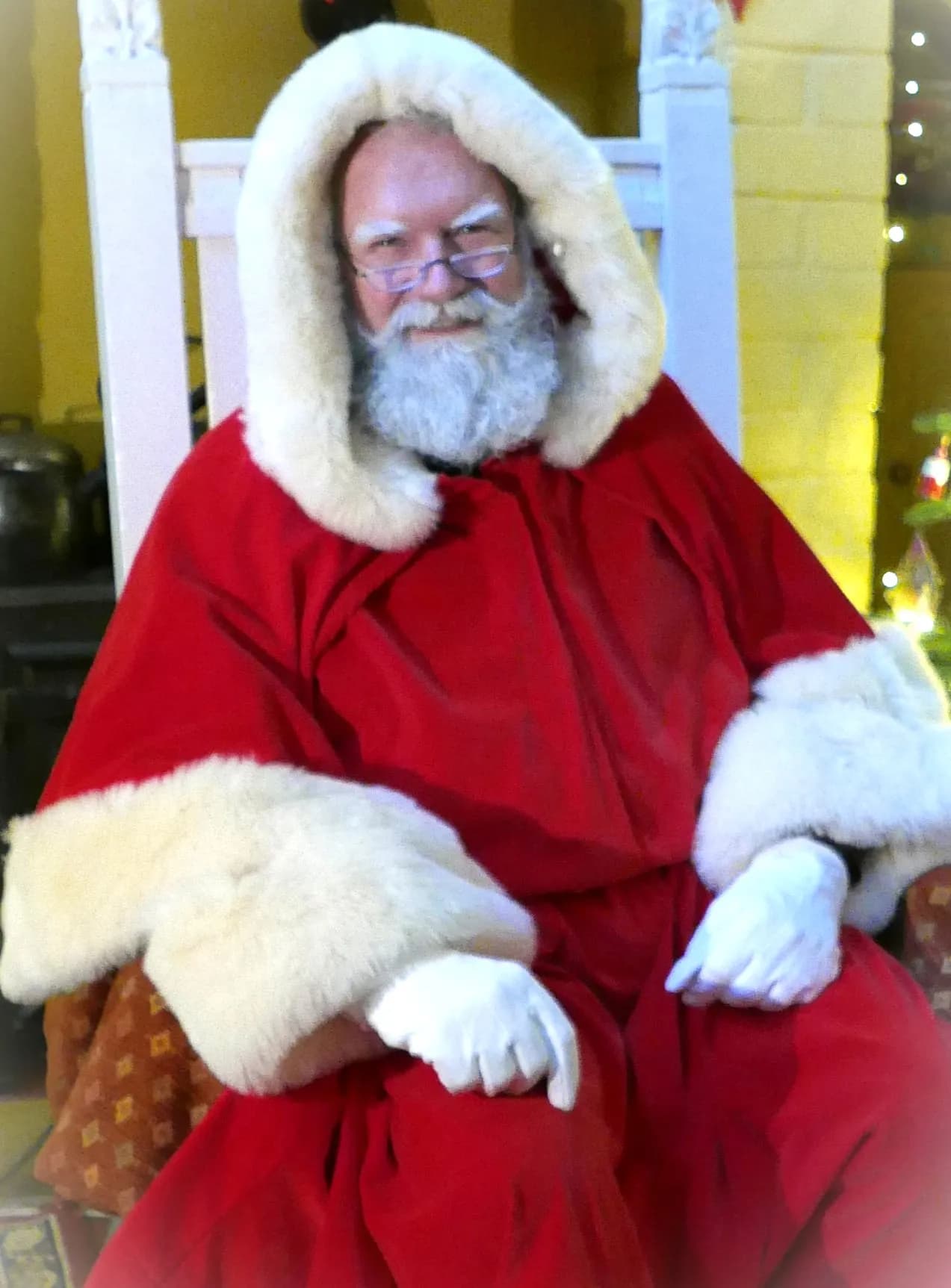 A person dressed as Santa Claus, wearing a red suit with white fur trim and white gloves, sits smiling in a chair.