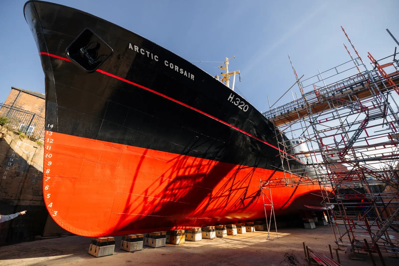 The bow of the "Arctic Corsair" trawler in a dry dock, with its black upper hull and red lower hull visible, surrounded by scaffolding.